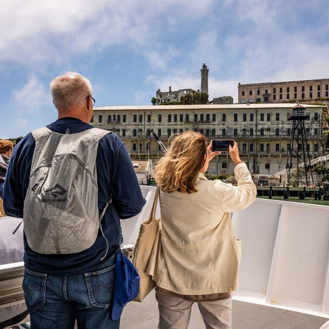 Two visitors on the bow of a boat, taking pictures of a large building on the Alcatraz dock.
