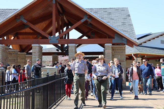 A group of people follow two NPS rangers as they walk away from a large stone structure.
