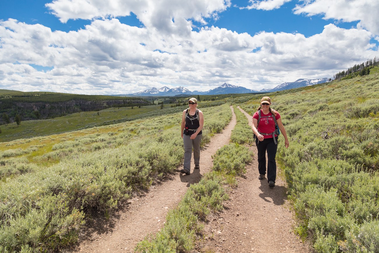 Two hikers on a trail surrounded by a grassy valley and mountains.