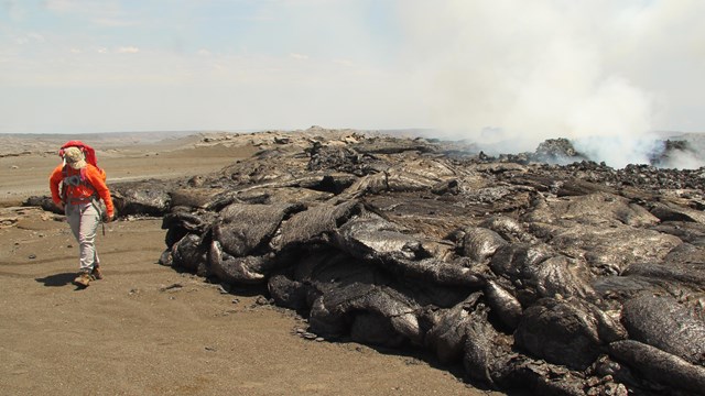 Figure wearing an orange safety shirt walking next to a new black cooling lava flow.